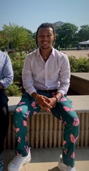 Photo of Alan Abutin sitting down outside and smiling.  He wears a white button down shirt and green pants with pink butterflies.