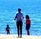 High contrast photo of a child on a beach with two adults; the water is a dazzling bright blue