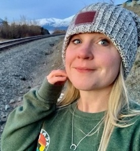 A blonde teenager smiles gently at the camera.  She is wearing a green sweatshirt and grey beanie.