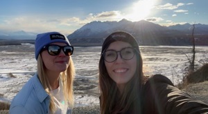 Two teenagers smile while taking a selfie.  There is snow and mountains in the background.  The one on the left is blonde, with a grey beanie and sunglasses, and the one on the right is a brunette with glasses and a black hat.