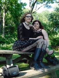 Kaelana and Penny sit together on a wooden picnic table, with green grass and trees in the background.  They both wear black.