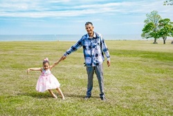 Photo of Olger and his daughter holding hands in the midst of green grass on a clear day.  He wears a blue flannel and she wears a pink dress, with a matching pink bow in her hair.  Both look cheerful.