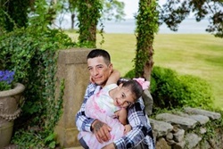 Photo of Olger in a gazebo with his daughter in his arms.  Both wear the same outfits as in previous photos.  Aynara has a huge smile and is missing a few baby teeth.