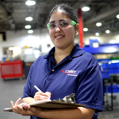 Young woman holding a clipboard. She is wearing safety glasses and a purple shirt. She is standing in a manufacturing facility.