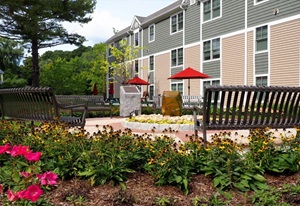 Image of a gray and beige apartment building with a park bench and flowers in the foreground