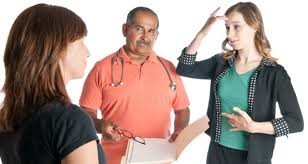 Three people in discussion.  A woman is using ASL to communicate with another woman while a man wearing a stethoscope looks on.