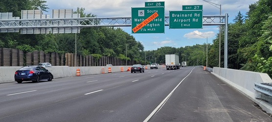 Picture from shoulder of highway, with Exit 28 sign overhead with a bright orange sign saying RAMP CLOSED covering the exit sign. Cars and trucks are driving underneath the sign.