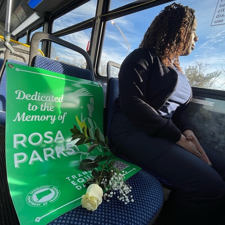 Person sitting on bus looking out the window. Next to them is a Transit Equity Day poster and rose, reserving a memorial seat in honor of Rosa Parks.