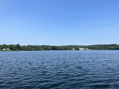 Landscape image of Chestnut Hill Reservoir. Blue sky with no clouds, green trees with hills and a few houses along the shoreline, and blue wavy water.