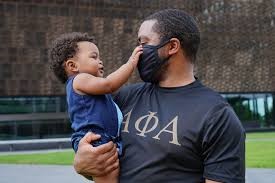 Jeffrey stands with in a dark grey fraternity t-shirt, holding his child