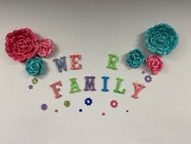 "We R Family" spelled in cutout letters with pink and turquoise paper flowers on a white background