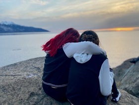 Photo of teens hugging each other in front of a body of water.  They face away from the camera.