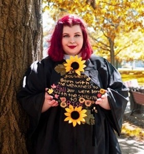 Kaelana in her graduation robes with her cap, which is decorated with sunflowers and the text "if you were born with the weakness to fall, you were born with the strength to rise."  She smiles gently and proudly.