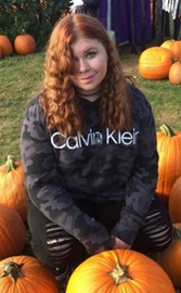 Photo of Kaelana smiling while sitting in a pumpkin patch.  Her bangs cover her right eye.