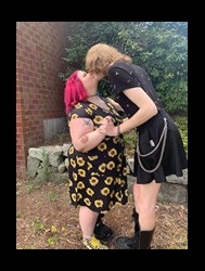 Photo of Kaelana kissing her partner Penny.  Kaelana wears a black sunflower dress and Penny is leaning over, wearing a black dress with a thin metal chain.