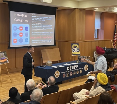 A man in a suit is presenting at a meeting in a conference room in Darien, Connecticut. A large screen displays information on ‘Hate/Bias Categories,’ and a table with DESPP branding is visible behind him. Several attendees are seated in the audience.