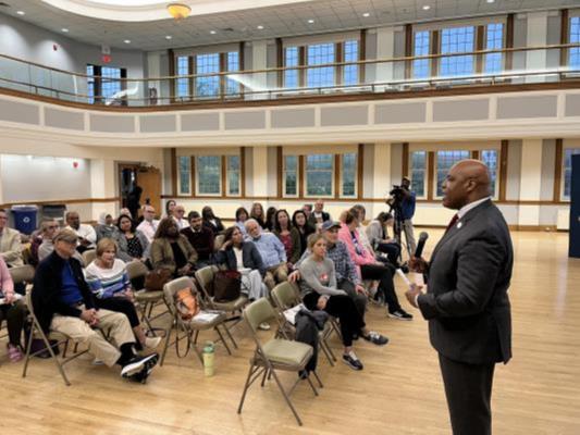 Commissioner Ronnell Higgins delivers remarks in a bright, open hall with high ceilings and large windows. A crowd of people listens attentively.