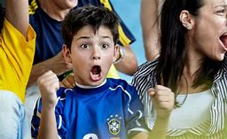 A young boy in a blue sports jersey cheers with his mouth open and fist raised while adults next to him cheer at a game.