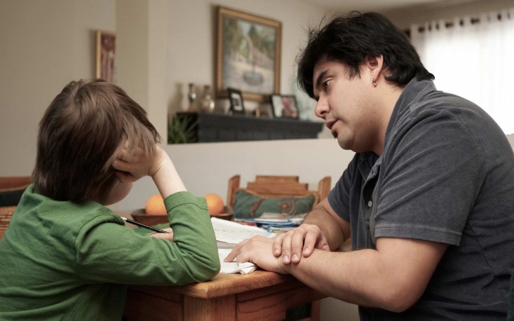 Dad sitting with a young child at a kitchen table having a serious discussion. 