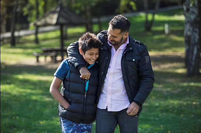 Father and son walking together outside with the father's arm around his son. Both are smiling and laughing.