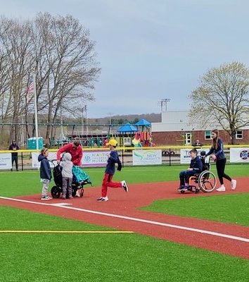 Children walking and running the bases on the accessible Miracle League field, including a student in a wheelchair pushed by a helper