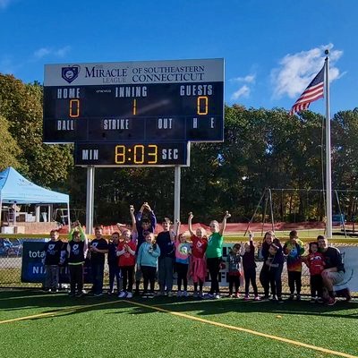 Smiling children with their arms raised in front of the Miracle League of Southeastern Connecticut scoreboard
