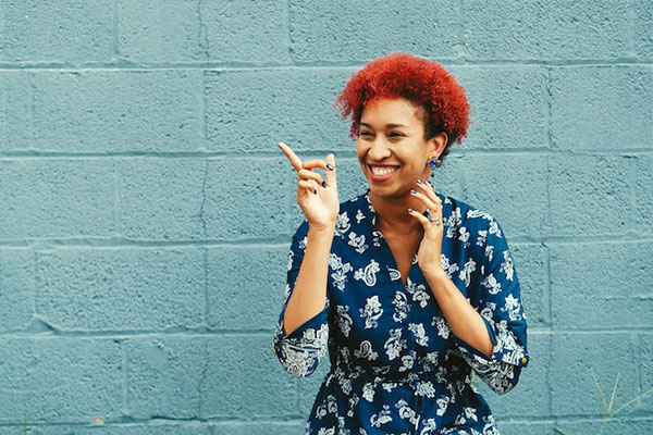 A black woman with colorful red hair smiles in front of a blue brick wall and points while looking in the distance