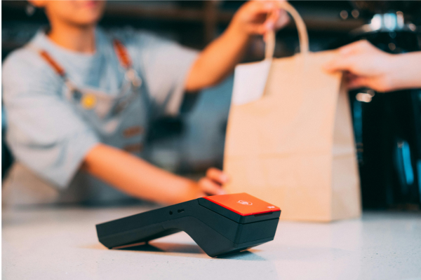 Image of cashier handing a shopping bag to a customer in the background. A contactless payment machine is placed in the foreground