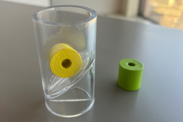 A small yellow bead about one inch in diameter sits inside a clear plastic test tube used to test small parts. A green bead is next to the tube.