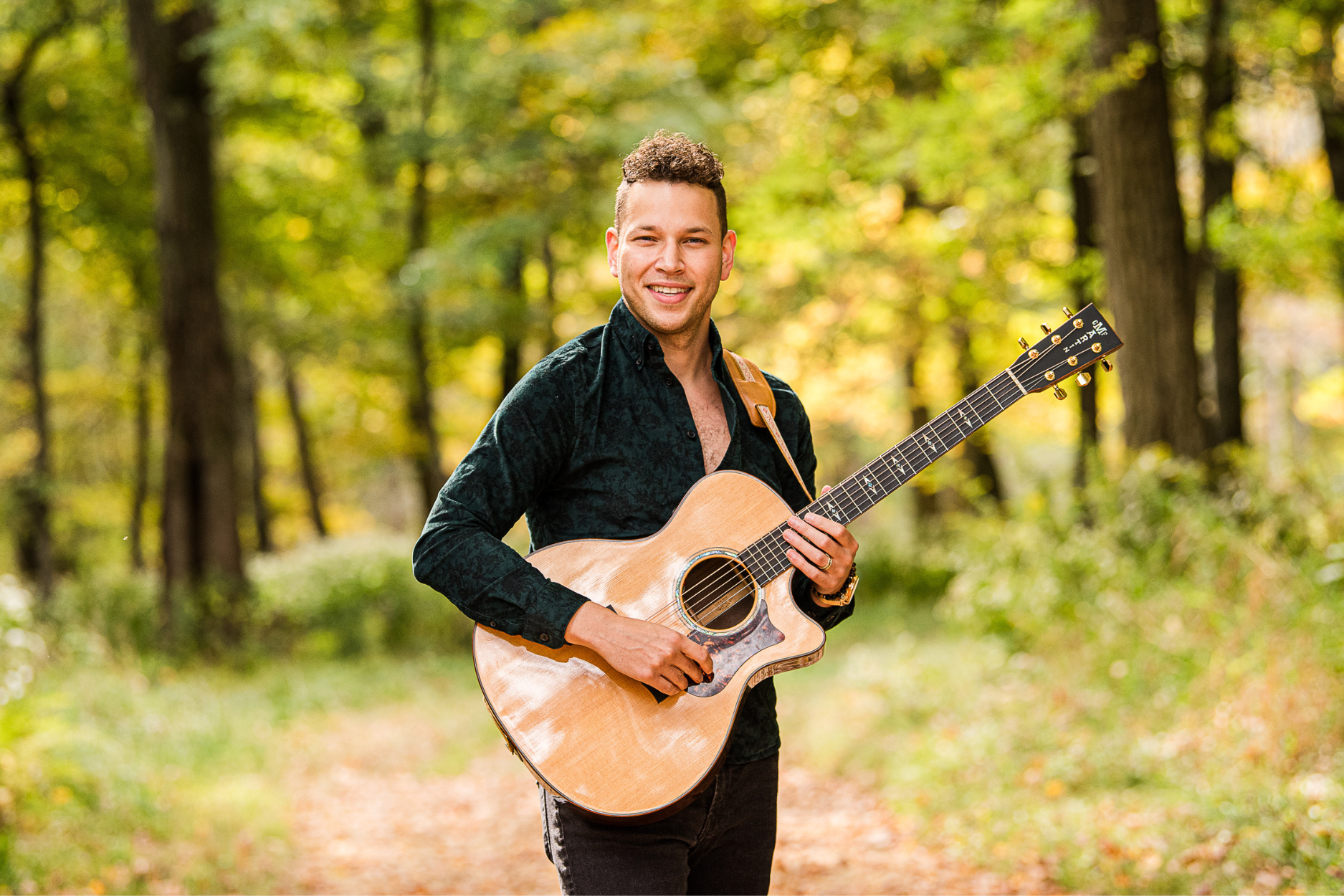 A person standing in a wooded area on a sunny day. The individual is holding an acoustic guitar with a light wood finish and dark tuning pegs. They are wearing a dark green, textured long-sleeve shirt and dark pants.  