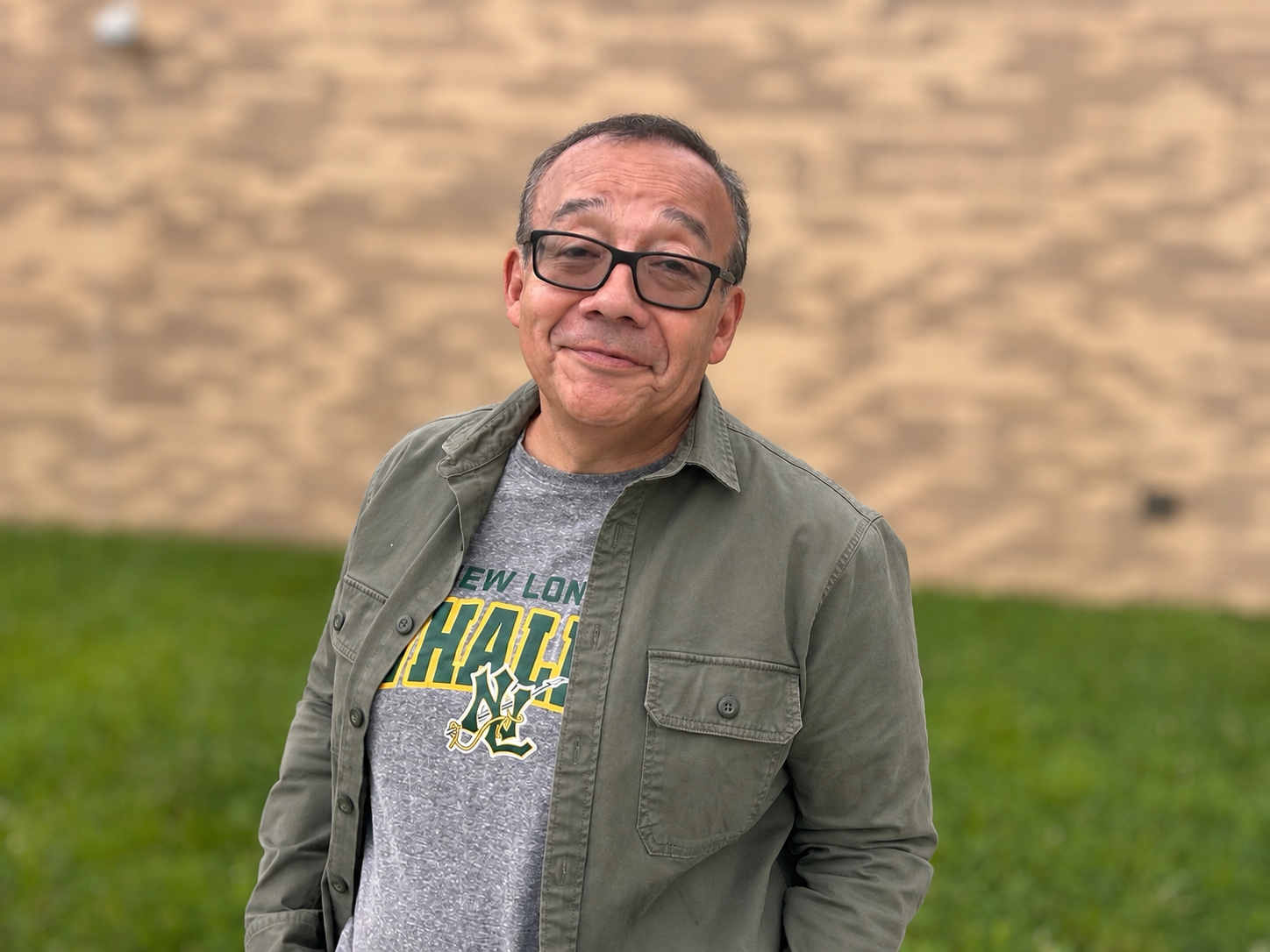 A man with glasses, short dark hair, and a slight smile stands in a grassy area in front of a blurred textured brick wall. 