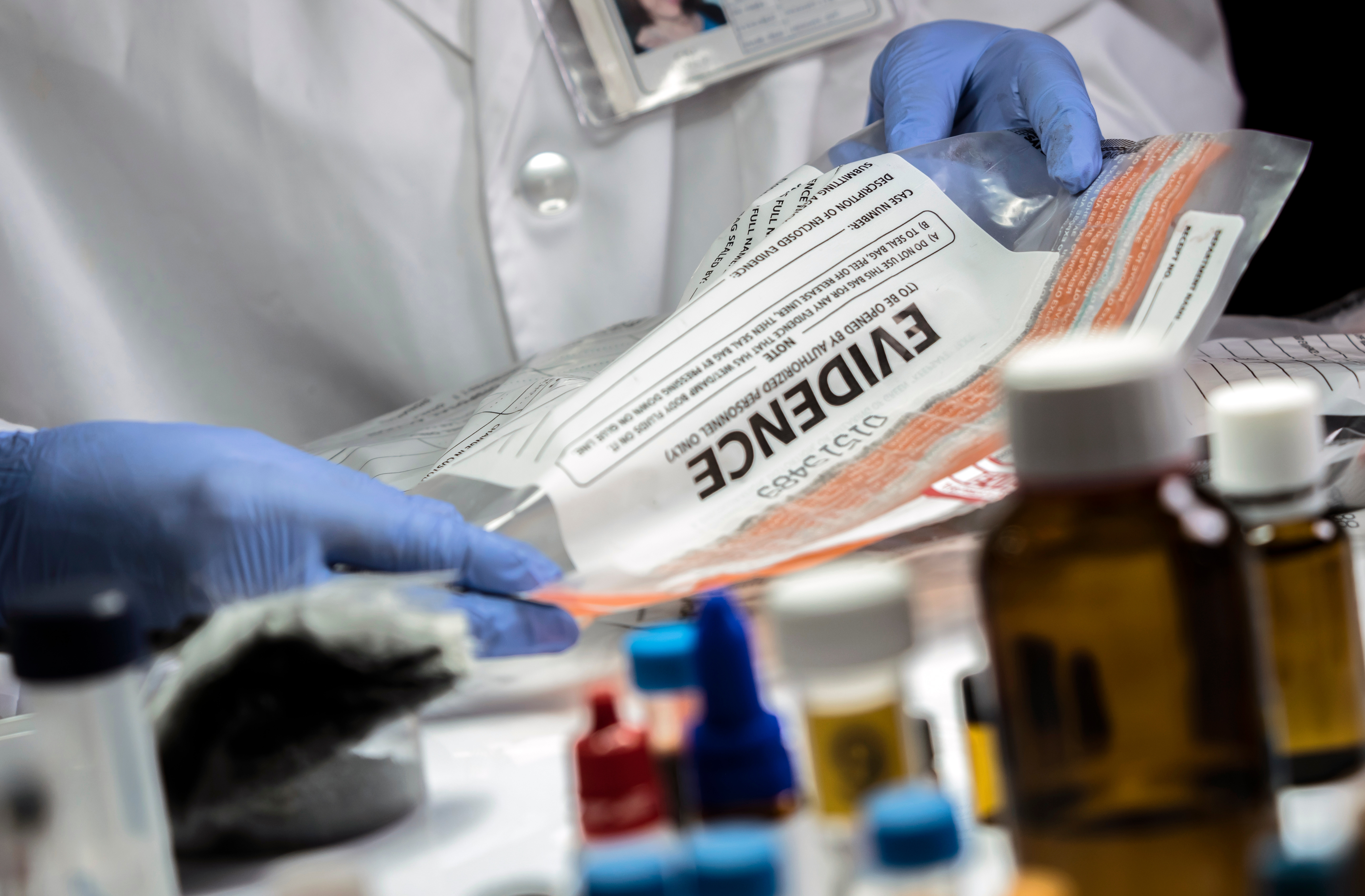 Forensic scientist wearing blue gloves examining an evidence bag in a laboratory, surrounded by chemical bottles and forensic tools
