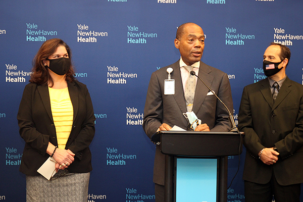 Commissioner and Yale New Haven Health staff members speaking behind podium with step and repeat banner.