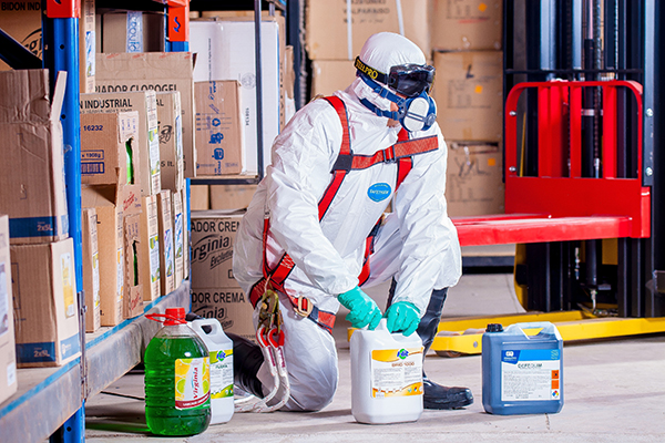 Person in protective work gear in chemical suit preparing cleaning solutions in a warehouse or industrial setting.
