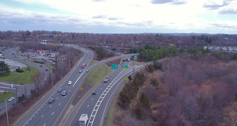 Aerial view of I-84 in Danbury, Connecticut, showing multiple lanes of traffic approaching Exit 3, surrounded by wooded areas and commercial areas