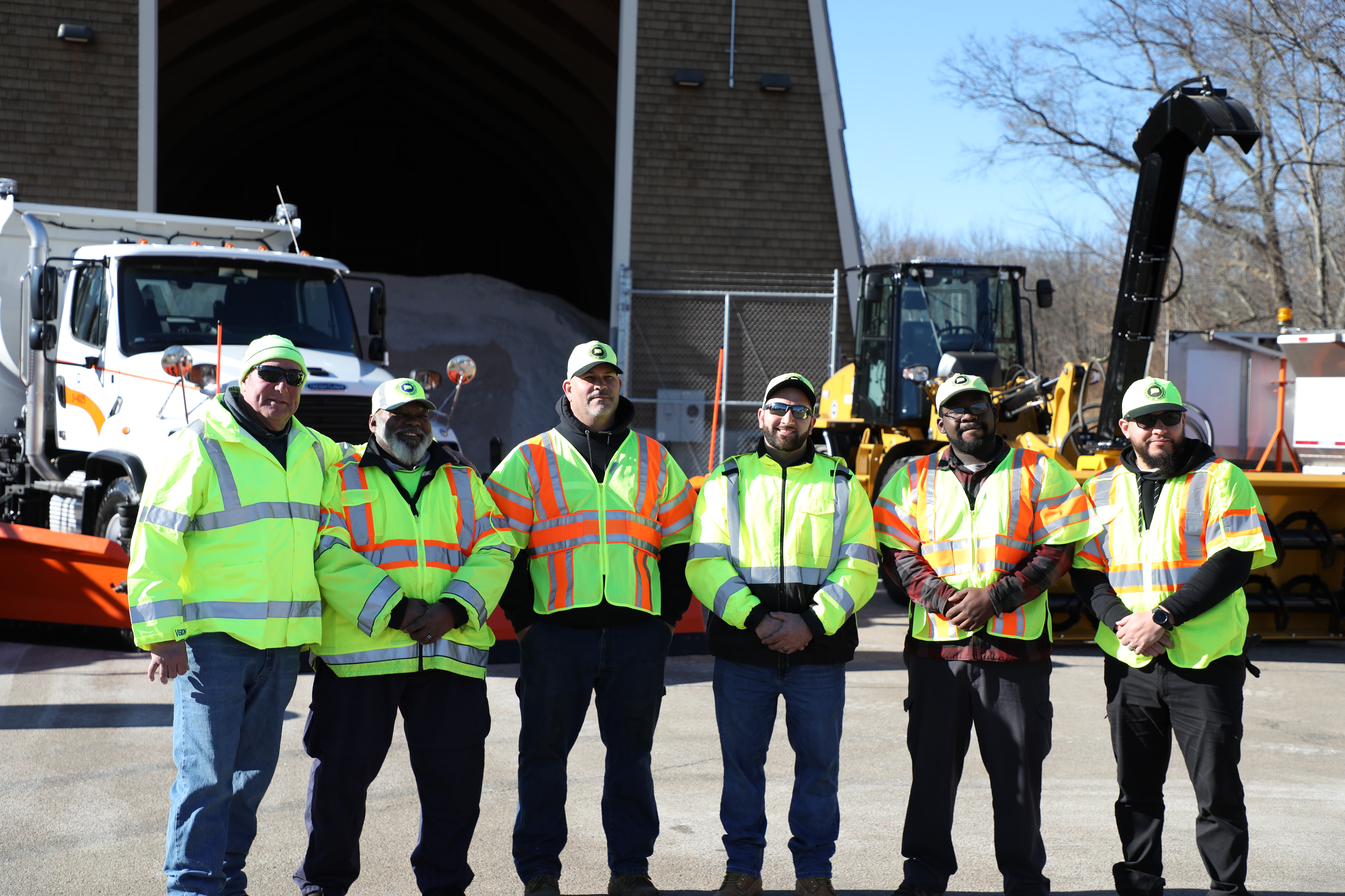 Photo of six men wearing yellow and orange reflective jackets and yellow hats with the CTDOT logo standing in front of construction equipment.