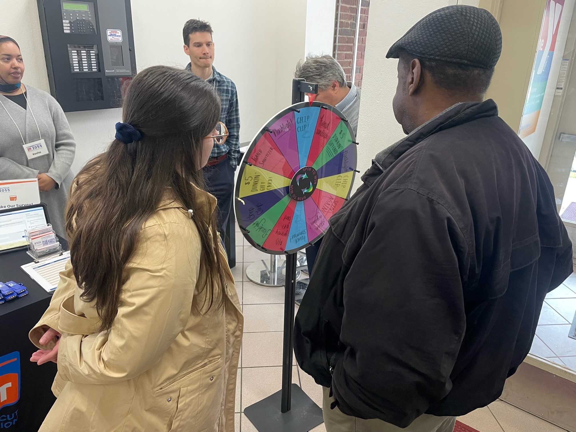 Photo of a woman wearing a camel colored jacket with long brown hair and man in a black coat and fedora style black hat facing a prize wheel at a pop-up event
