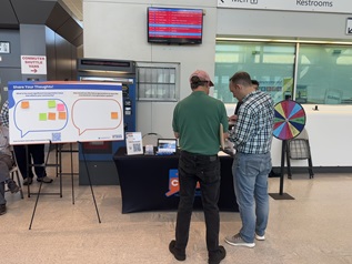 Image of two men standing at a pop up. The man to the left is wearing a red hat, green polo shirt, and black pants. The man to the right is wearing a blue and white plaid shirt and jeans. Both men are facing away to the camera with their heads down looking at something. A board with sticky notes and a prize wheel can be seen in the back.