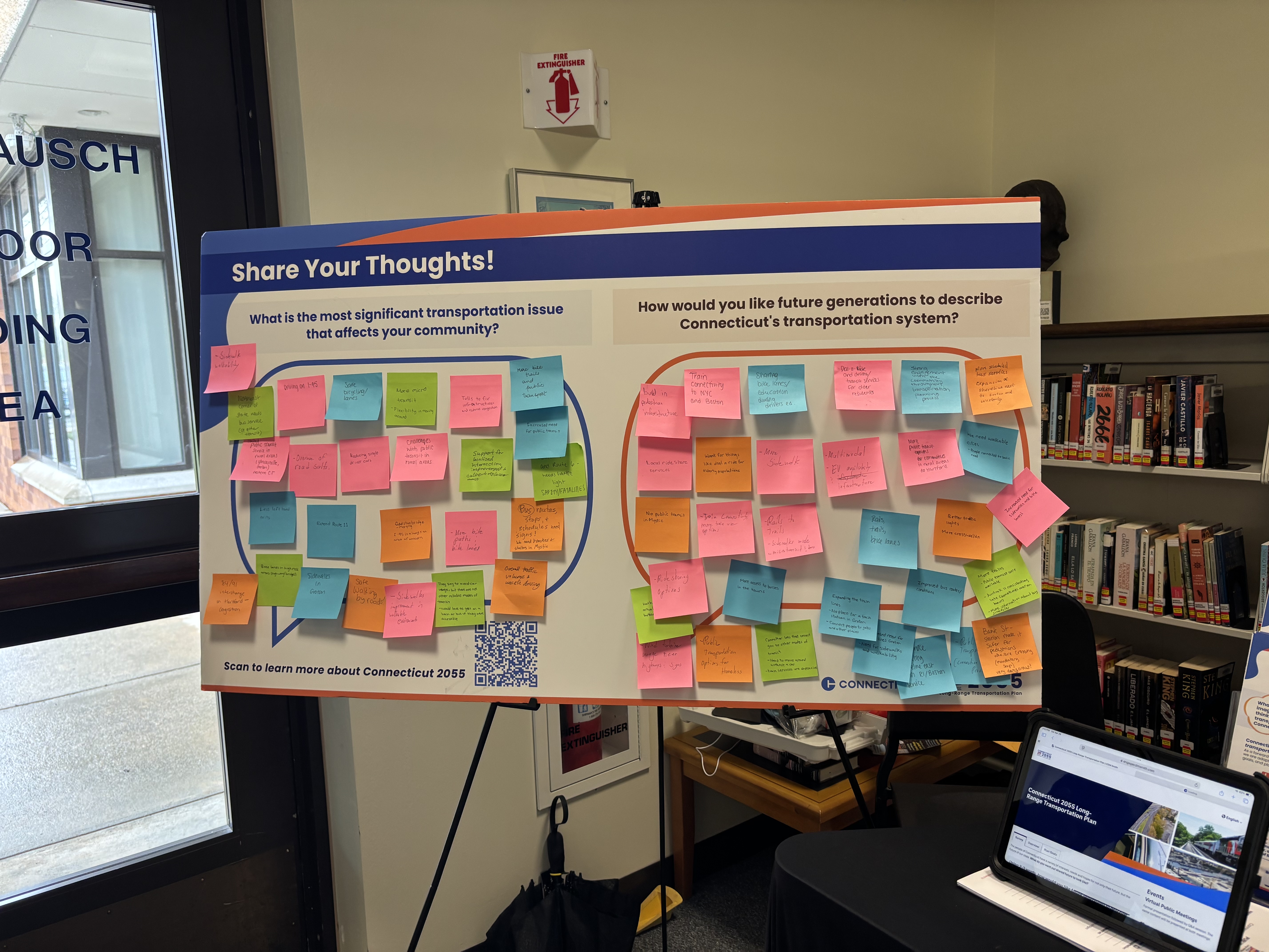 Photo of a board on an easel. The board has a blue and orange border and is titled "Share Your Thoughts". There are multiple different colored sticky notes covering the board. 