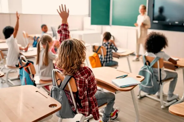 A group of students sitting at desks in a classroom with a teacher standing at the front of the room.