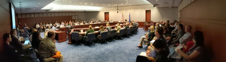 A photograph of the PFAS Task Force Meeting held on August 28, 2019. Task Force members are seated around a meeting table, while members of the public, state agency staff, and other interested parties observer from seats along the site of the room. 