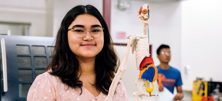 female student stands next to a model of a skeleton
