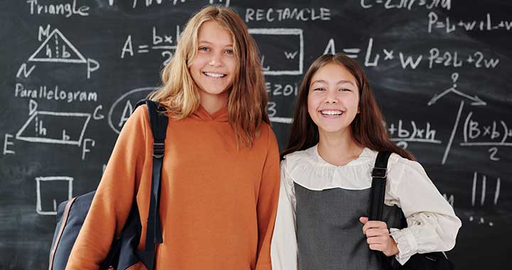 two pre-teen girls with backpacks stand in front of a chalkboard